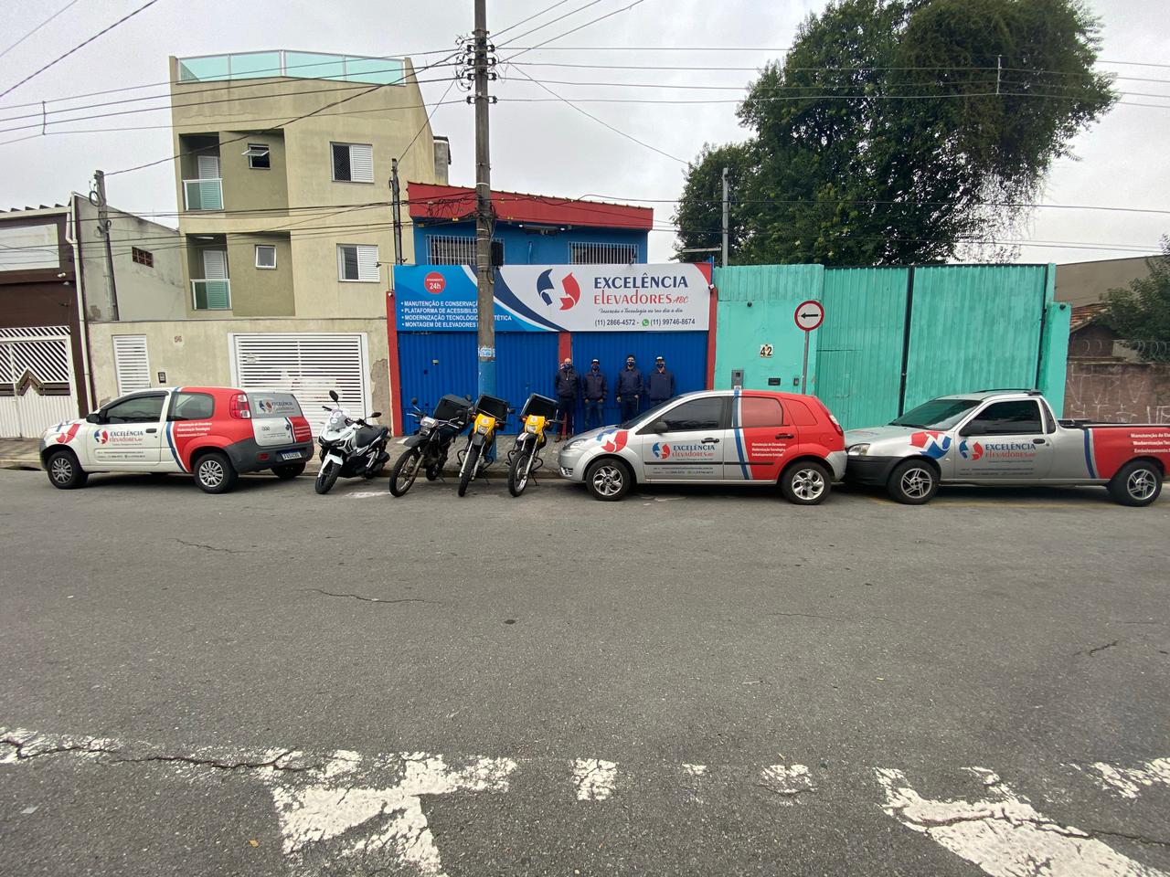 Técnicos uniformizados da Excelência Elevadores ao lado dos carros, da picape e das motos da empresa, na porta da sede